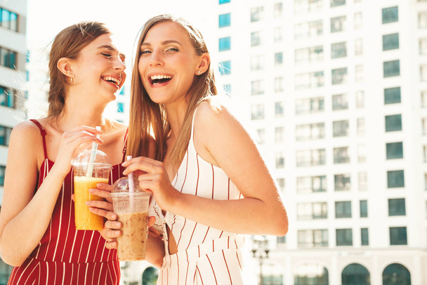 Two young beautiful smiling hipster female in trendy summer clothes. Carefree women posing outdoors.Positive models holding and drinking fresh cocktail smoothie drink in plastic cup with straw - Photo, image
