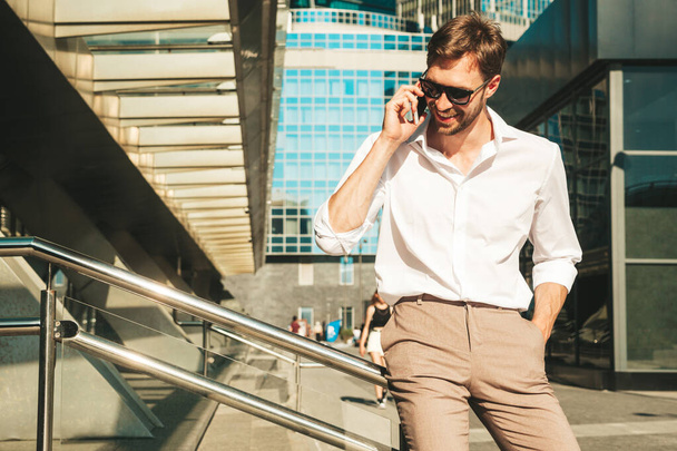 Handsome smiling stylish hipster lambersexual model.Modern man dressed in white shirt. Fashion male posing on the street background in sunglasses. Outdoors at sunset. Using smartphone.Talking - Φωτογραφία, εικόνα
