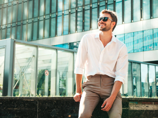 Portrait of handsome smiling stylish hipster lambersexual model.Modern man dressed in white shirt. Fashion male posing in the street background near skyscrapers in sunglasses. Outdoors at sunset  - Fotografie, Obrázek