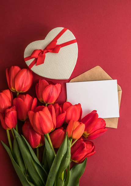 赤いチューリップの花束とギフトのハートボックスは赤い背景に隔離されています。春の花。誕生日、女性、母の日、結婚式、バレンタインデーのグリーティングカード。平置きだ。コピースペース。バナー - 写真・画像