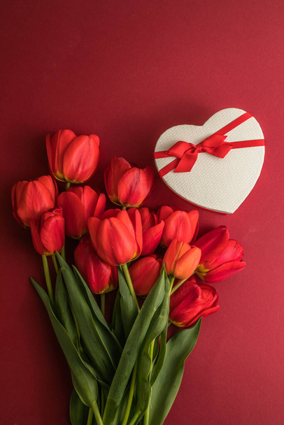 Kimppu punainen tulppaanit ja lahja sydän laatikko eristetty punaisella pohjalla. Kevätkukkia. Äitienpäivä, Häät, Syntymäpäivä, Ystävänpäivä, Nainen. Litteä pano. Kopioi tilaa. Banneri - Valokuva, kuva