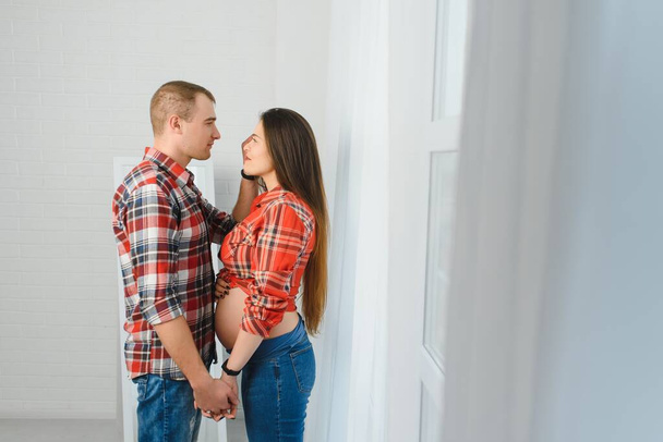 Loving man hugging his pregnant wife from behind standing near window at home, copy space - Photo, Image