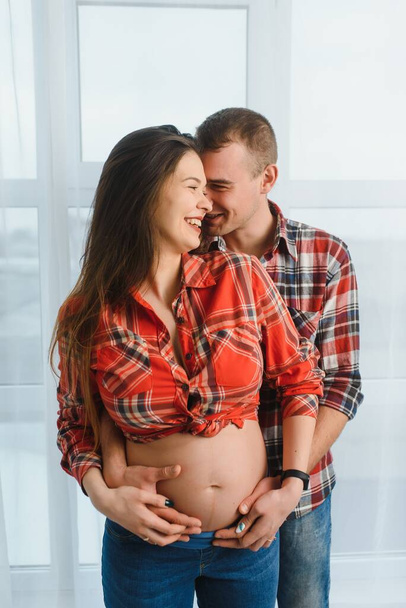 Loving man hugging his pregnant wife from behind standing near window at home, copy space - Zdjęcie, obraz