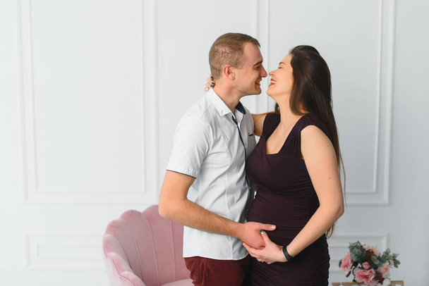 Smiling pregnant young couple hugging while standing together at a window - Foto, Bild