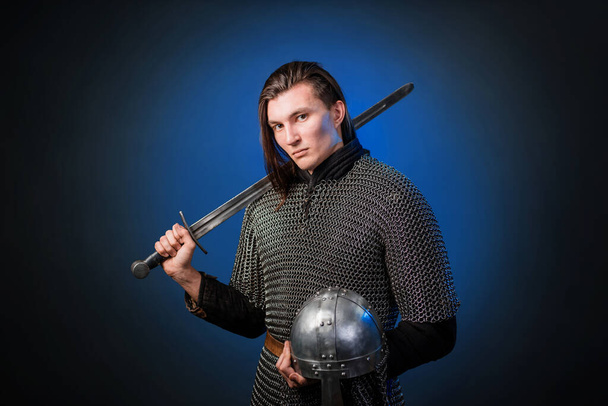Portrait of a medieval warrior of the late Viking era and the beginning of the Crusades. Knight with long hair in chain mail, armed with a sword isolated on a dark, black and blue background. - Photo, Image