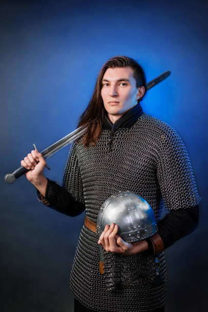 Portrait of a medieval warrior of the late Viking era and the beginning of the Crusades. Knight with long hair in chain mail, armed with a sword isolated on a dark, black and blue background. - Foto, afbeelding