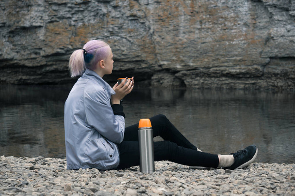 Teenager-Wanderin ruht sich am Ufer des Flusses mit einem Becher aus und blickt auf die Felsen an der Küste - Foto, Bild
