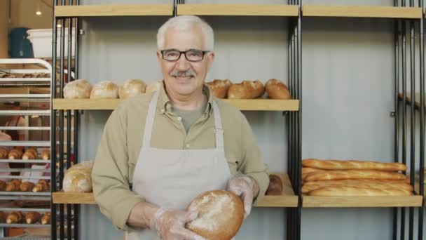 Tracking portret z powolnym tempie szczęśliwy starszy mężczyzna piekarz w fartuchu i rękawice trzymając świeżo pieczony chleb i uśmiechając się do kamery w jego przytulnej piekarni - Materiał filmowy, wideo