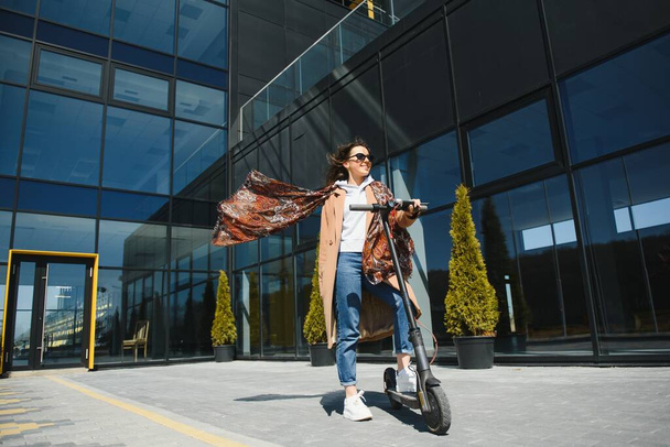 A young beautiful girl is standing next to her electric scooter. She is smiling, business-trained, with modern architecture in the background. - Photo, image