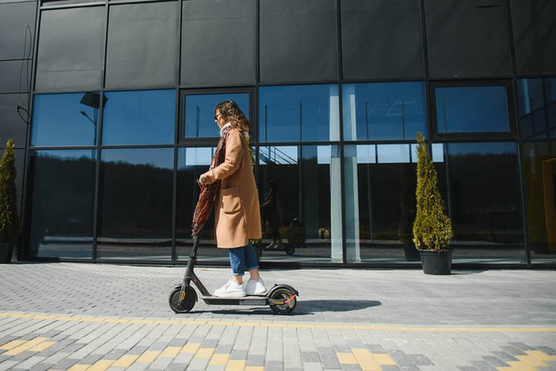 A young beautiful girl is standing next to her electric scooter. She is smiling, business-trained, with modern architecture in the background. - Foto, afbeelding