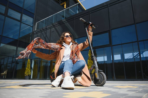 Beautiful young woman in sunglasses is sitting on her electro scooter near big glass building. - Foto, imagen