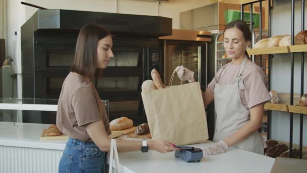 Slowmo shot of young female customer using mobile phone to pay for paper bag full of freshly baked goods in cozy bakery Happy female counter attendant in apron smiling and thanking customer - Footage, Video