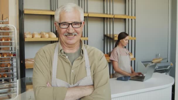 Slowmo portrait shot with tracking of happy elderly man in apron and glasses standing in cozy bakery and smiling for camera Female counter attendant working on laptop in background - Footage, Video