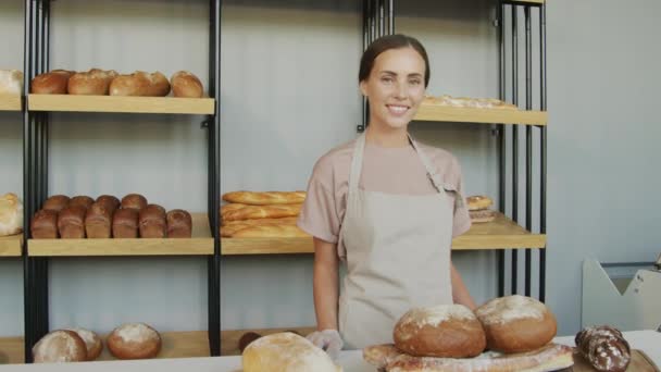Rastreamento retrato tiro com lentidão de jovem assistente de balcão feliz posando na padaria aconchegante com pães recém-assados em prateleiras - Filmagem, Vídeo