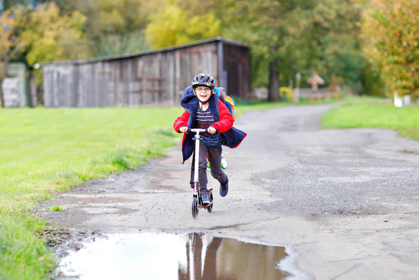 милый маленький школьник, катающийся на скутере по дороге в школу или из школы. Школьник 7 лет ехал по дождевой луже. Смешной счастливый ребенок в красочной одежде и в шлеме. - Фото, изображение