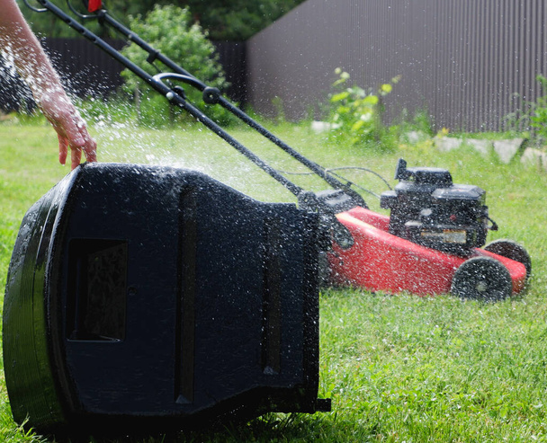 Washing the lawn mower's grass catcher with water. Splashes of water on a background of green grass. A gas-powered self-propelled lawn mower in the background. - Photo, Image