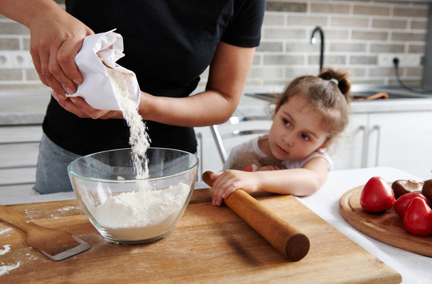 Kobieta wlewająca białą mąkę z papieru do szklanej miski. Obok niej siedzi jej córeczka i ogląda proces gotowania. - Zdjęcie, obraz