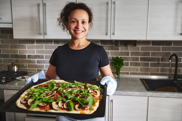 Mutfak eldivenlerinde gülümseyen İspanyol kadın fırından sıcak pizzayla fırın örtüsü tutuyor, ev mutfağının arka planında duruyor. - Fotoğraf, Görsel