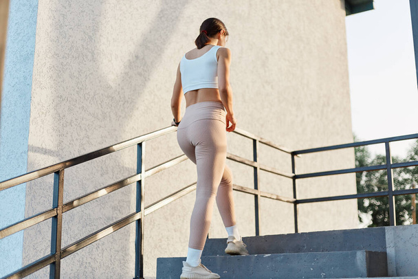 Back view of slim female with good shape wearing stylish sporty clothing, beige leggings and top, going upstairs training outdoor, healthy lifestyle, lady training. - Foto, Bild