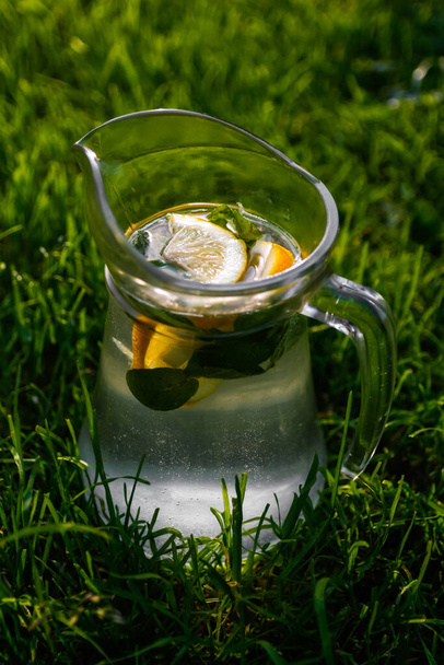 Defokussieren Sie Nahaufnahme Glas Krug Limonade mit Zitrone und Minze auf natürliche grüne Natur Hintergrund. Krug mit kaltem Sommercocktail oder Punsch im Freien, draußen. Trinken Sie Mojito. Vertikal. Unscharf. - Foto, Bild