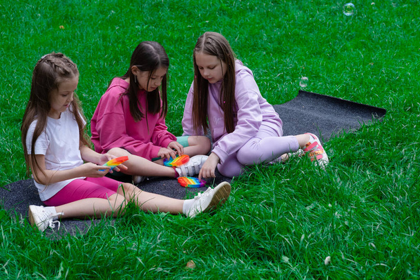 tři dospívající dívky si hrají s módní smyslovou hračkou pop to venku na trávě. Push pop bublina flexibilní pohyblivé pohyblivé hračka. Stres a úleva od úzkosti. Módní silikonová hra pro stresované děti a dospělé. - Fotografie, Obrázek