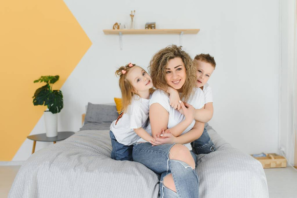 Portrait of smiling young mommy embracing kids little son and daughter. Happy family. Mother with children sitting on bed and loving the good times together. Closeup - Fotografie, Obrázek