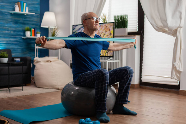 Jubilado hombre mayor sentado en la pelota suiza en la sala de estar haciendo ejercicios de fitness de salud - Foto, Imagen