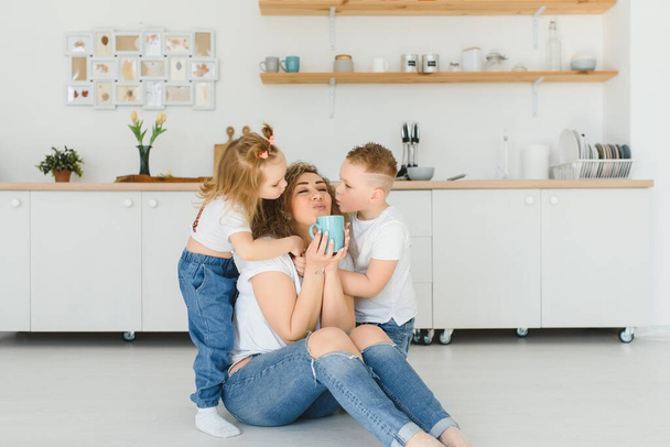 Happy mother hugging daughter and son sitting on wooden floor in modern kitchen at home. Mom hugging children. Happy family relationship concept - Foto, afbeelding