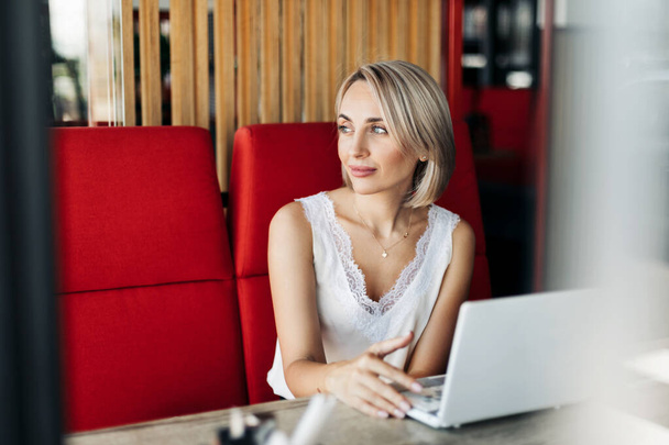 Une belle blonde utilise un ordinateur portable et travaille dans un café, regarde attentivement au loin. - Photo, image