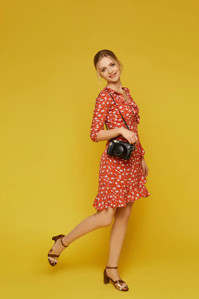Full-length portrait of a model girl in a colorful summer dress posing with a vintage camera over yellow background, isolated. Young caucasian woman in a trendy summer dress on a yellow background - Photo, image