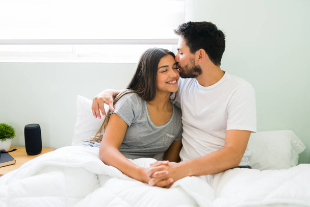 Красивая девушка и парень в возрасте 20 лет целуются и улыбаются после пробуждения в постели в солнечное утро в день отдыха - Фото, изображение