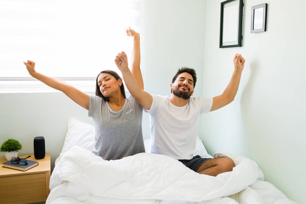 Τι όμορφη μέρα! Ήρεμη νεαρή γυναίκα και άντρας τεντώνουν τα χέρια τους ενώ νιώθουν ξεκούραστοι αφού ξυπνήσουν μαζί σε ένα άνετο κρεβάτι. - Φωτογραφία, εικόνα