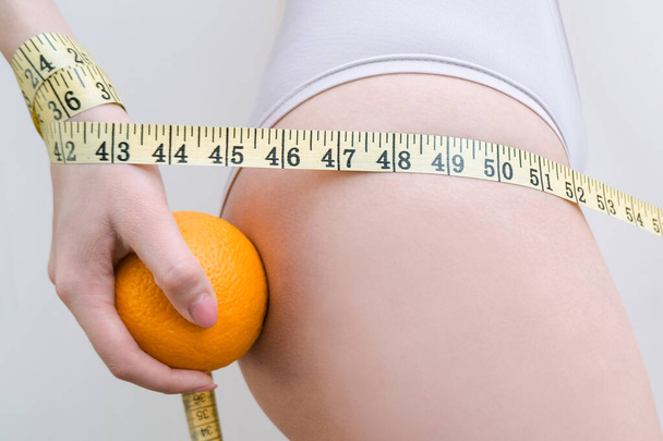 Młoda kobieta trzyma pomarańczę i taśmę pomiarową na jasnym tle. Koncepcja problemu cellulitu - Zdjęcie, obraz