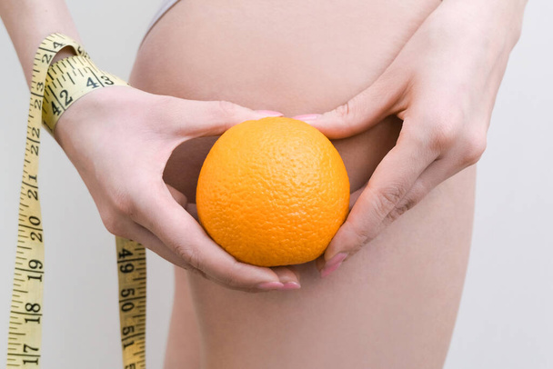Μια νεαρή γυναίκα κρατάει ένα πορτοκάλι και μια μεζούρα σε ελαφρύ φόντο. Κυτταρίτιδα έννοια πρόβλημα - Φωτογραφία, εικόνα