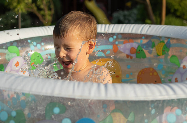 Mały chłopiec rozpryskuje się w nadmuchiwanym basenie. Zezulce od sprayu. Koncepcja wakacji na podwórku - Zdjęcie, obraz