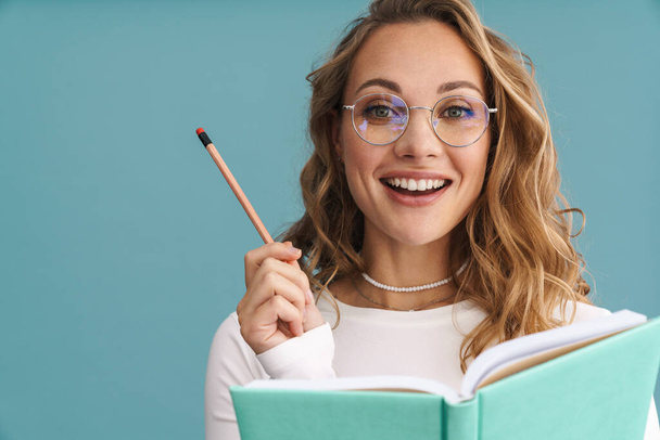 Smiling young woman in glasses holding pencil and book over blue wall background - Valokuva, kuva