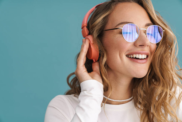 Smiling young blonde woman in glasses listening to music with wireless headphones over blue wall - Foto, Imagen