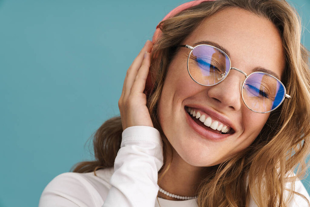 Smiling young blonde woman in glasses listening to music with wireless headphones over blue wall - Zdjęcie, obraz