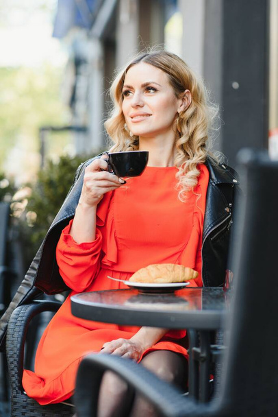 Młoda kobieta pije kawę w ulicznej kawiarni. Pani z pięknym makijażem w drogiej bluzce jest tajemniczo. - Zdjęcie, obraz