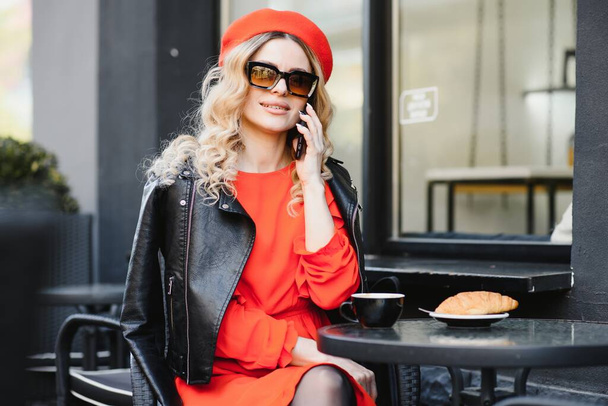 Fashionable woman talking by phone at outdoors cafe. Fashion and beauty. Technology and lifestyle concept. Stylish woman with perfect makeup and hairstyle. Seductive girl at coffee shop outside. - Photo, image