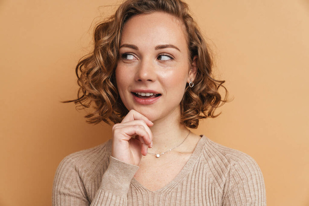 Young ginger woman with wavy hair smiling and looking aside isolated over beige background - Photo, Image