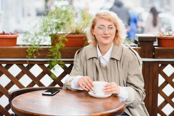 young stylish beautiful woman sitting in city cafe in street, spring fashion trend style, drinking latte, pink purse, exited, emotional, happy, smiling - Photo, Image