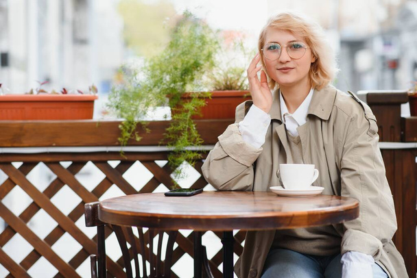 Very beautiful young woman, sit in Cafe and drink coffee or tea, street front view - Фото, зображення
