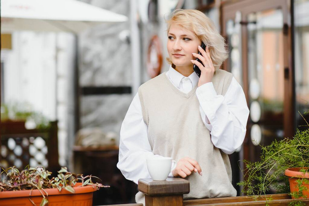 Fashionable woman talking by phone at outdoors cafe. Fashion and beauty. Technology and lifestyle concept. Stylish woman with perfect makeup and hairstyle. Seductive girl at coffee shop outside. - Foto, afbeelding