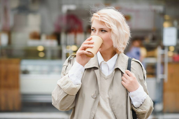 Cheerful stylish young woman in the street drinking morning coffee in sunshine light. - Photo, Image