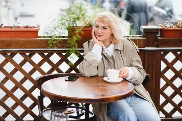 young stylish beautiful woman sitting in city cafe in street, spring fashion trend style, drinking latte, pink purse, exited, emotional, happy, smiling - Photo, Image