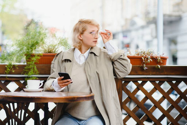 young stylish beautiful woman sitting in city cafe in street, spring fashion trend style, drinking latte, pink purse, exited, emotional, happy, smiling - Foto, Imagem