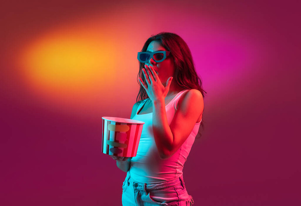 Excited in 3D eyewear and with popcorn. Caucasian womans portrait isolated on red studio background in pink neon light. Beautiful male model. - Photo, Image