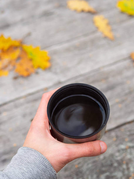 Puchar termosu z gorącą herbatą w ręku na drewnianym stole z żółtymi jesiennymi liśćmi. Herbata grzewcza w podróży - Zdjęcie, obraz