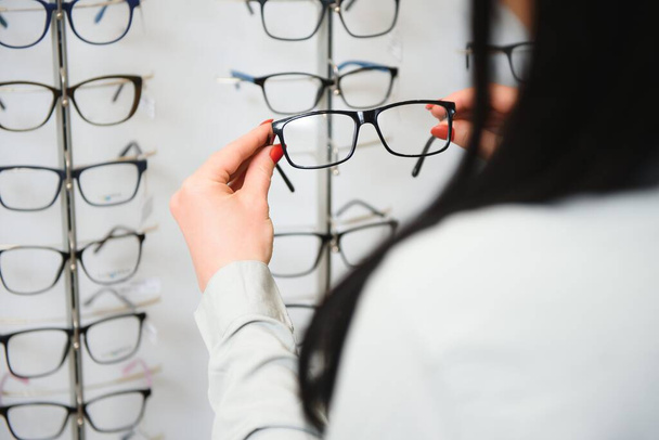 Row of glasses at an opticians. Eyeglasses shop. Stand with glasses in the store of optics. Woman's hand chooses spectacles. Eyesight correction. - Фото, изображение